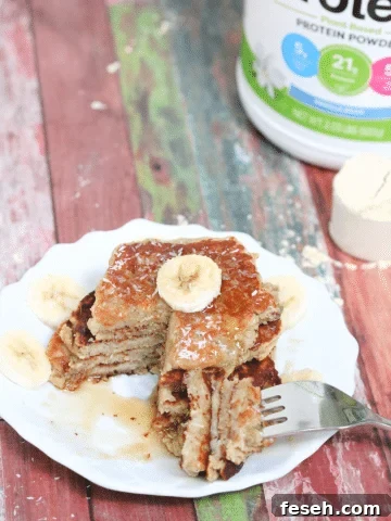 White plate with banana protein pancakes stacked and a slice cut out and on the fork beside the place, banana on and maple syrup top of the pancakes and protein powder behind them