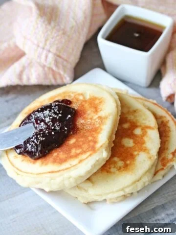 Three yogurt pancakes on a white plate topped with jam. Syrup in a white container behind it and a light orange napkin