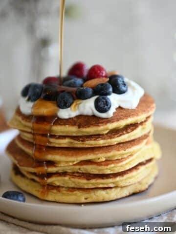 Stacked protein pancakes topped with whipped cream and berries and syrup being poured on them.