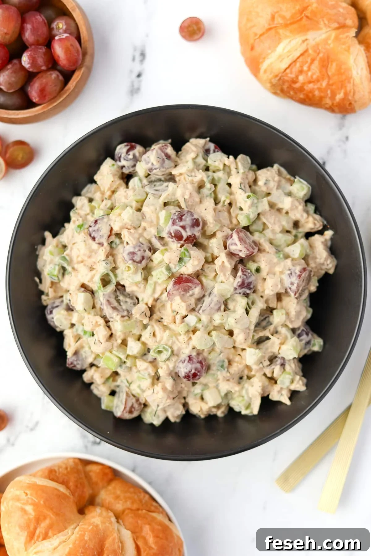 A bowl of chicken salad in a black bowl, surrounded by a bowl of grapes and some croissants. 