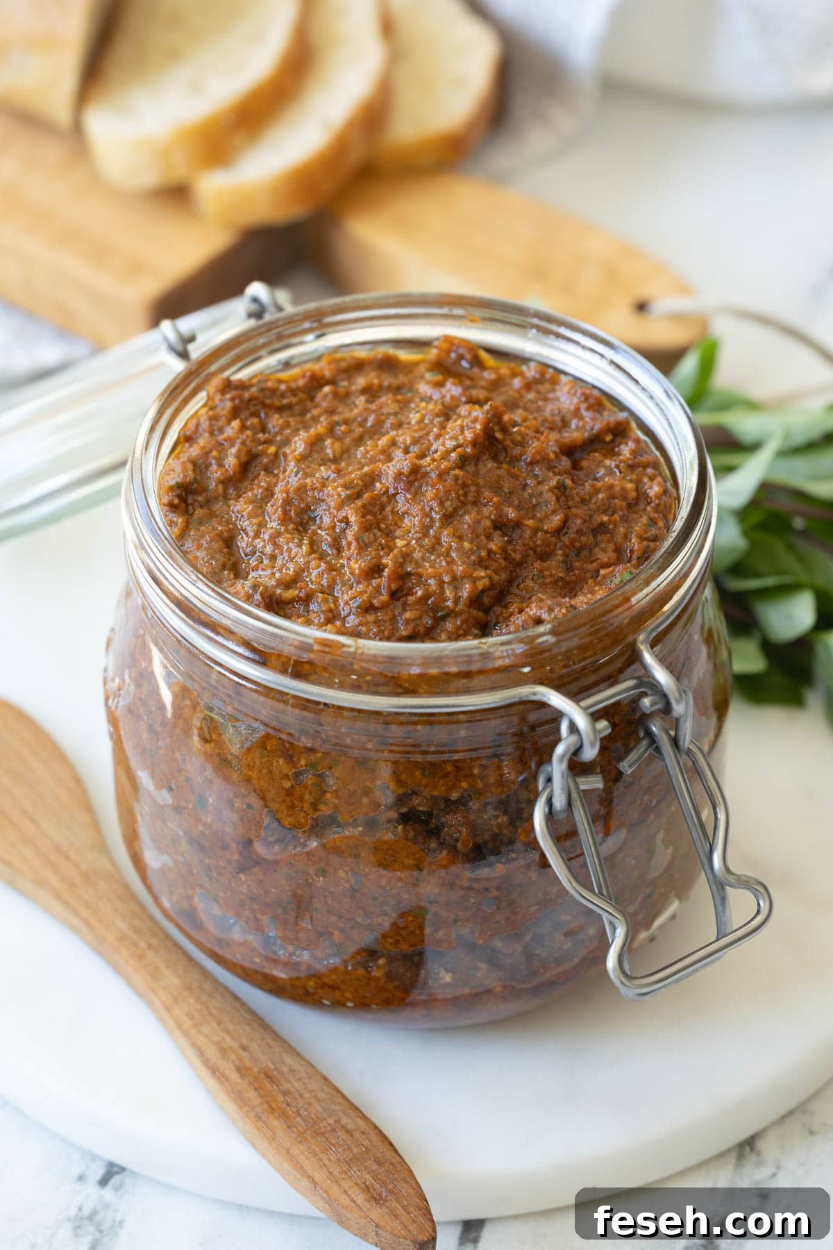 An open glass jar of sun-dried tomato pesto with bread slices behind and basil leaves.