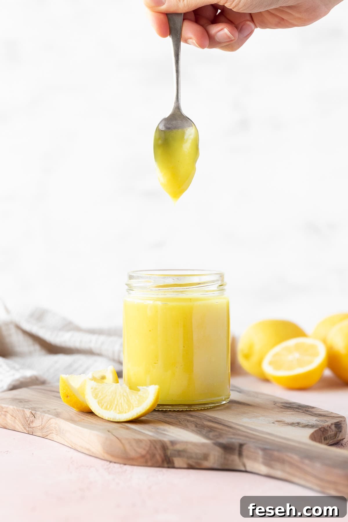 Side on shot of a hand holding a spoon of vegan lemon curd vertically over the full mason jar with the curd slowly dripping off.