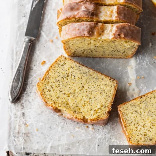 A slice of gluten free lemon poppy seed cake laying on parchment paper with a knife next to it and the sliced loaf behind it.