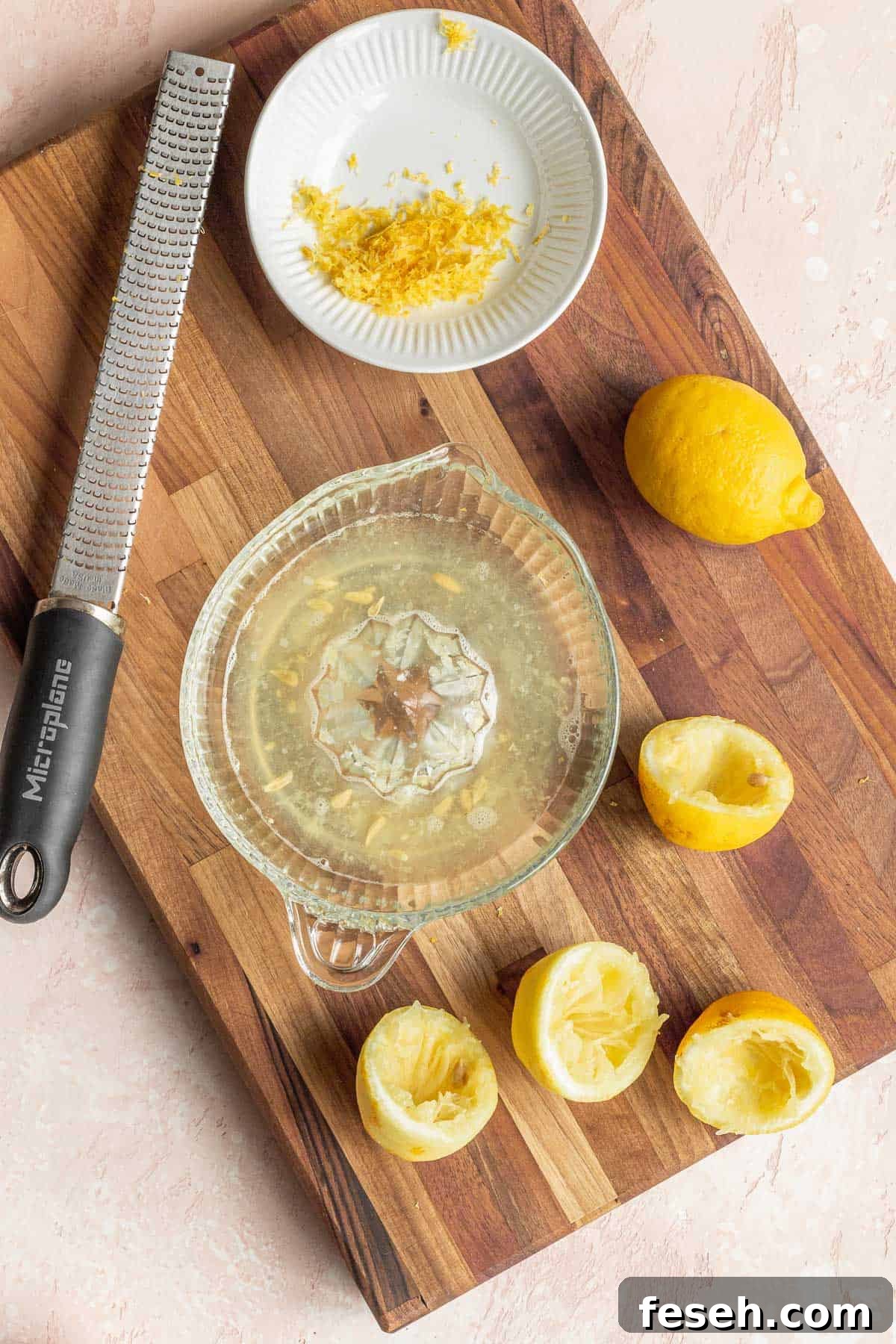 overhead shot of an old fashioned manual lemon juicer bowl filled with juice, a microplane, a plate of lemon zest, and several spent lemons on a wooden cutting board.