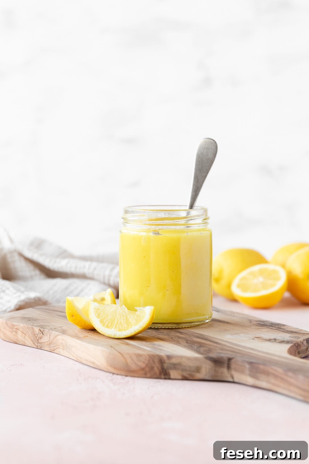 Side on shot of a small mason jar of vegan lemon curd with a silver spoon in it on a wooden cutting board with a few wedges of fresh lemon.