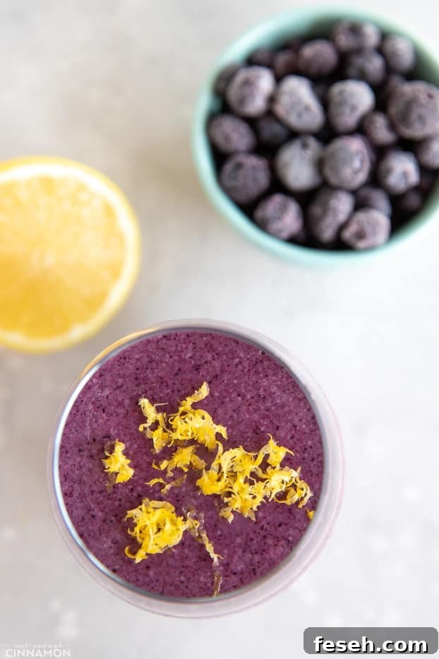 Lemon blueberry smoothie topped with lemon zest, with frozen blueberries and half a lemon on the side