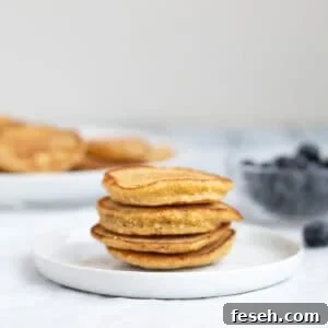 4 pancakes stacked on a white plate with a bowl of blueberries behind