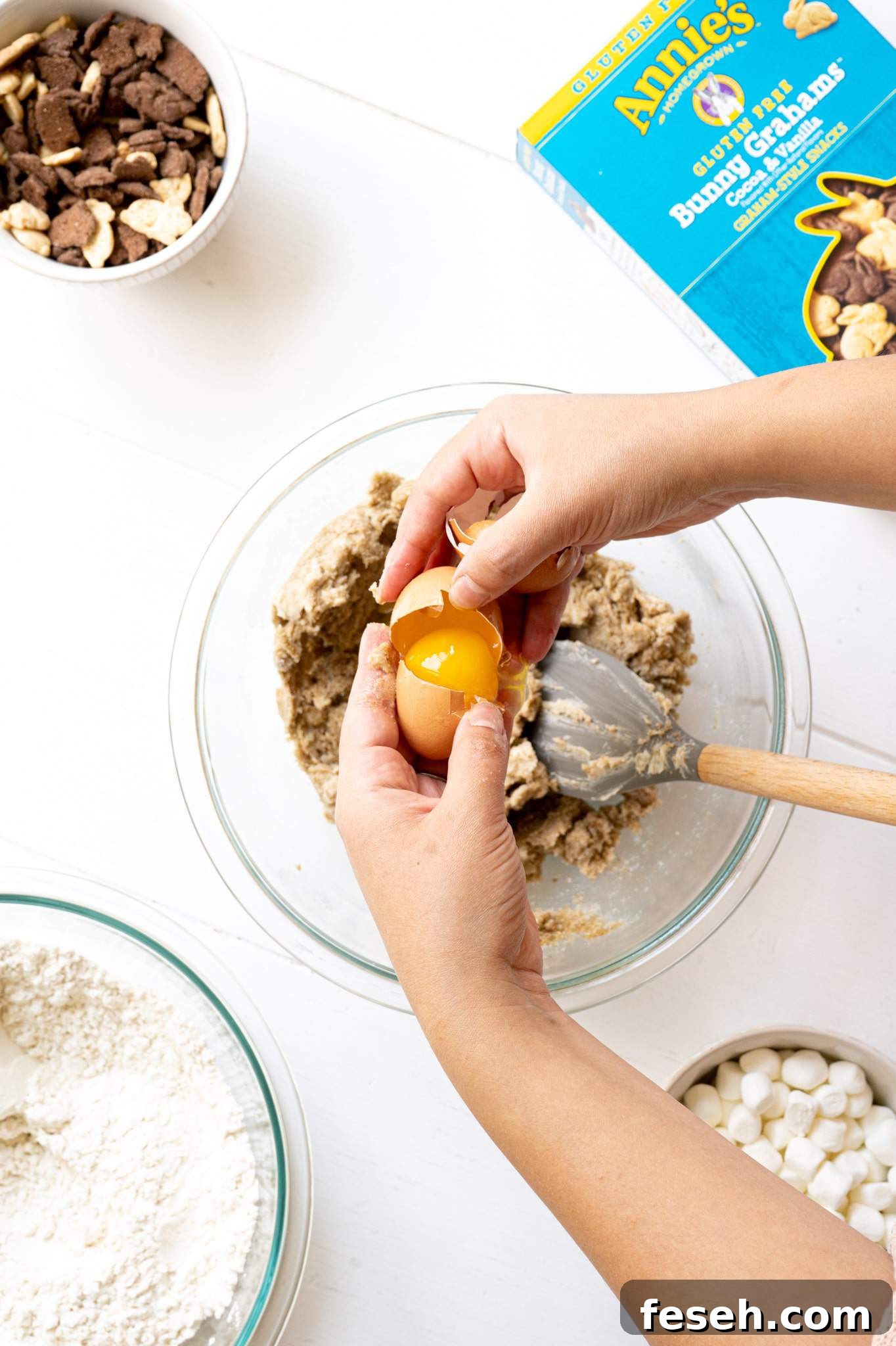 Two hands cracking an egg into s'mores cookie dough.