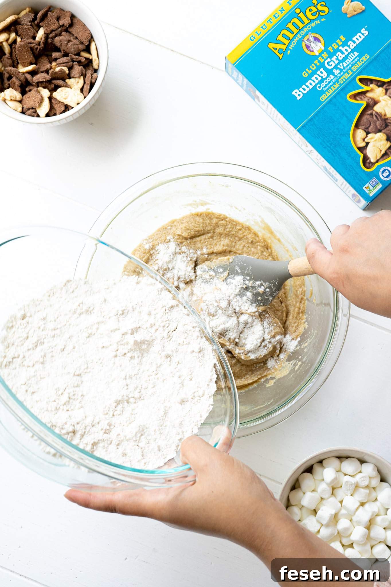 A hand pouring flour into a mixing bowl with butter, sugar, eggs and vanilla.