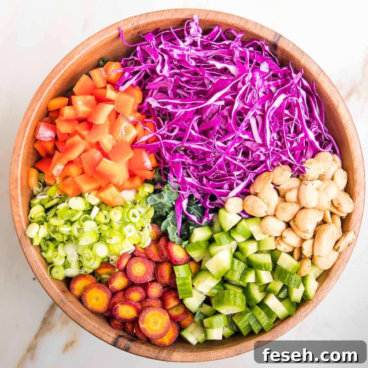 Wooden salad bowl filled with chopped kale and then sections of chopped veggies are on top of the kale.  Purple cabbage, red peppers, scallions, purple carrots, cucumbers and marcona almonds