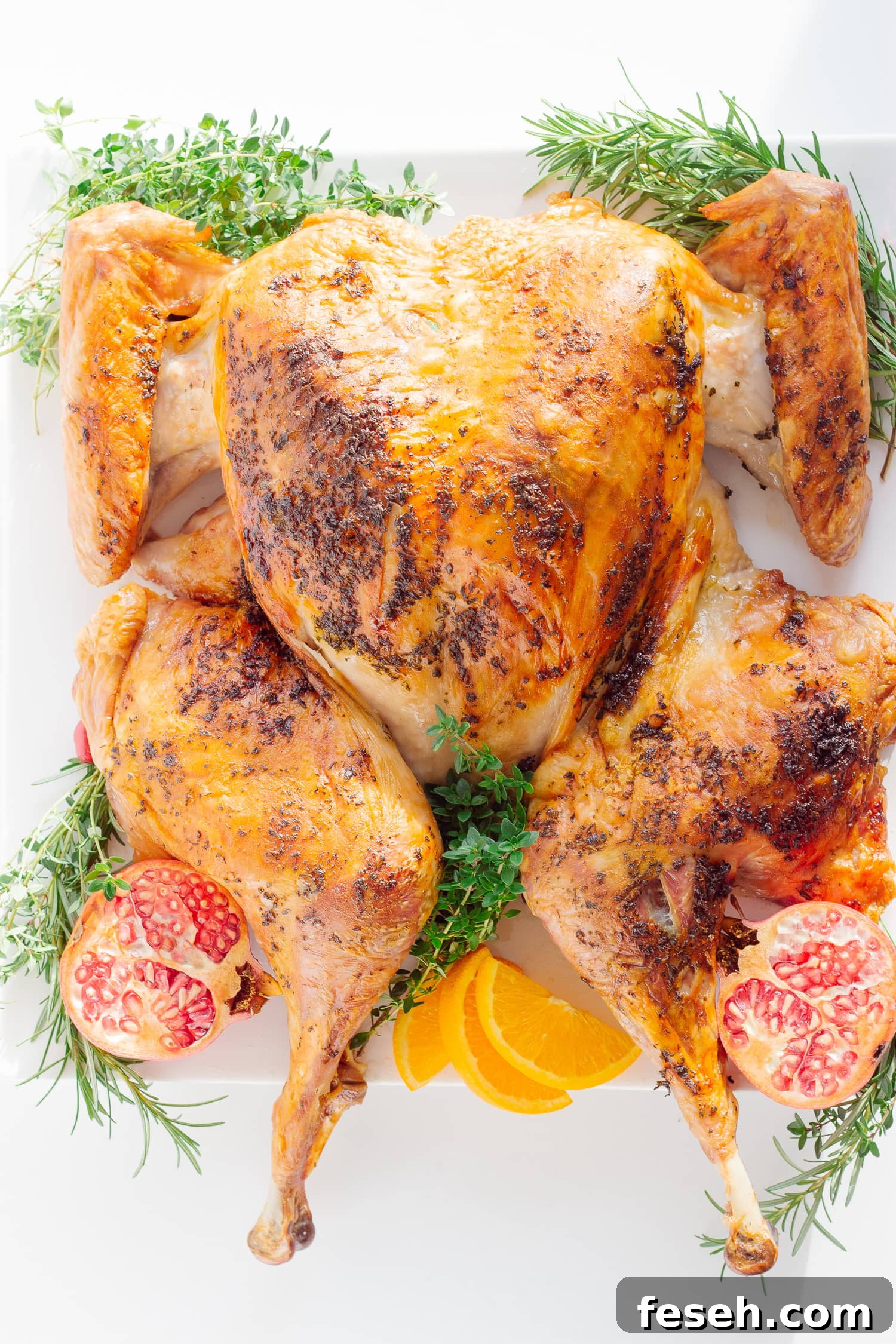 Roasted Spatchcock Turkey with herb butter on a platter with herbs, pomegranate and orange slices scattered around.