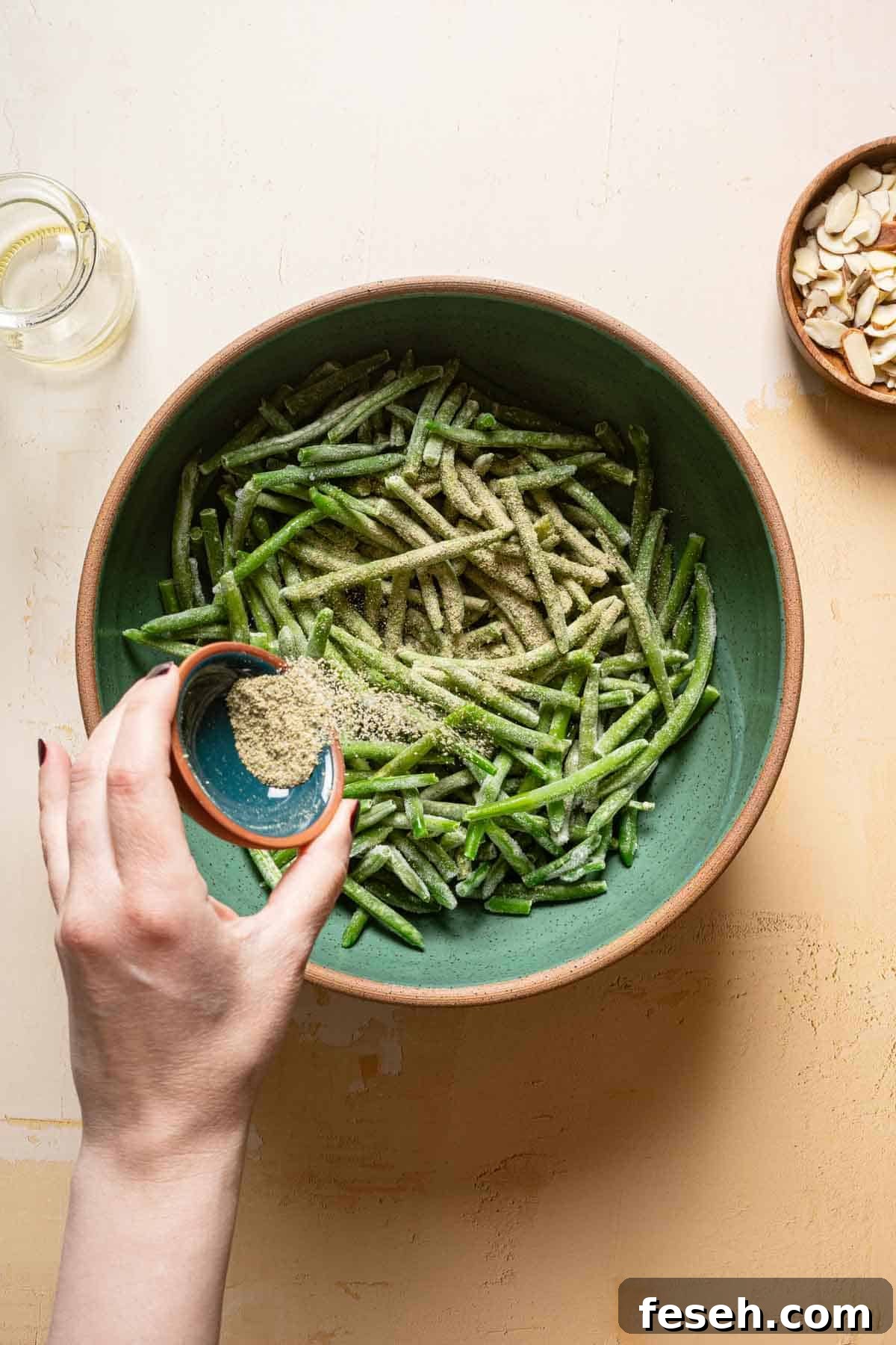 hand pouring spices overtop frozen green beans in bowl.