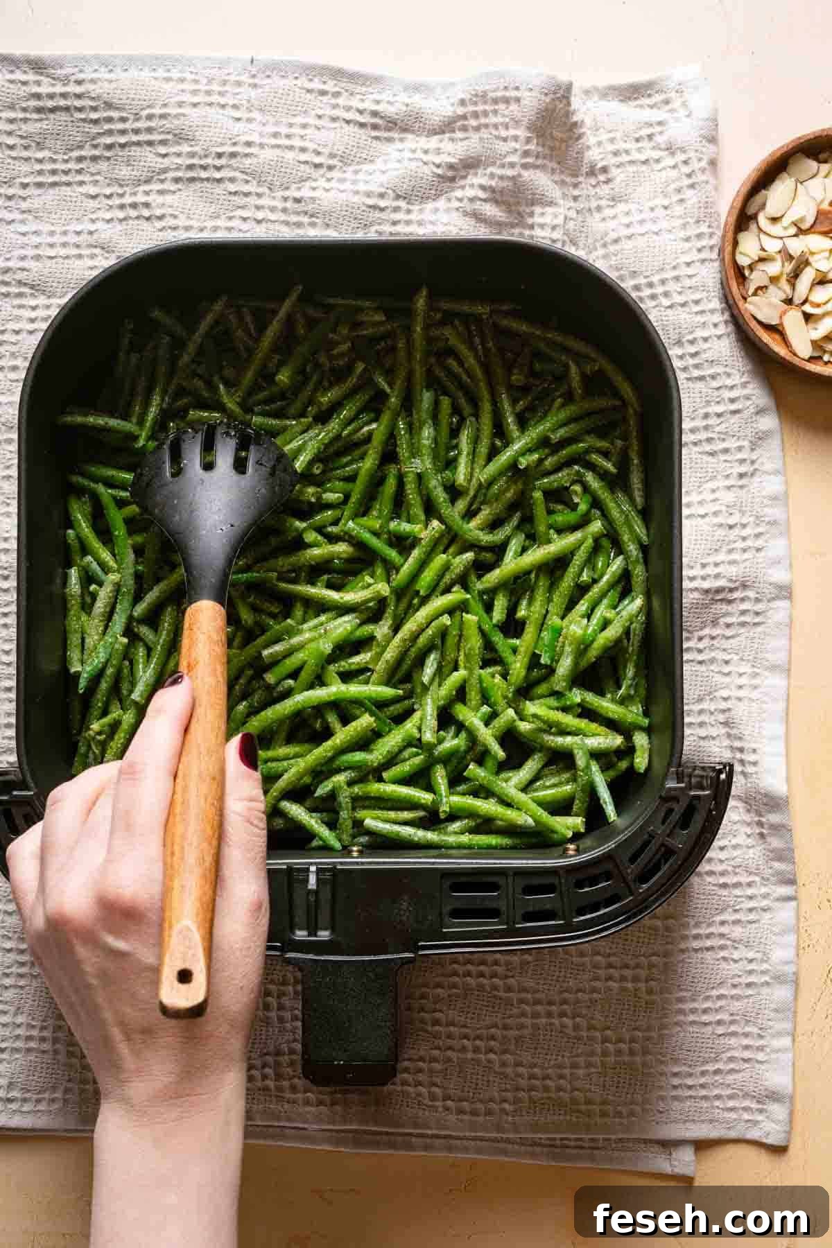 partially cooked green beans being stirred in air fryer basked with spoon.