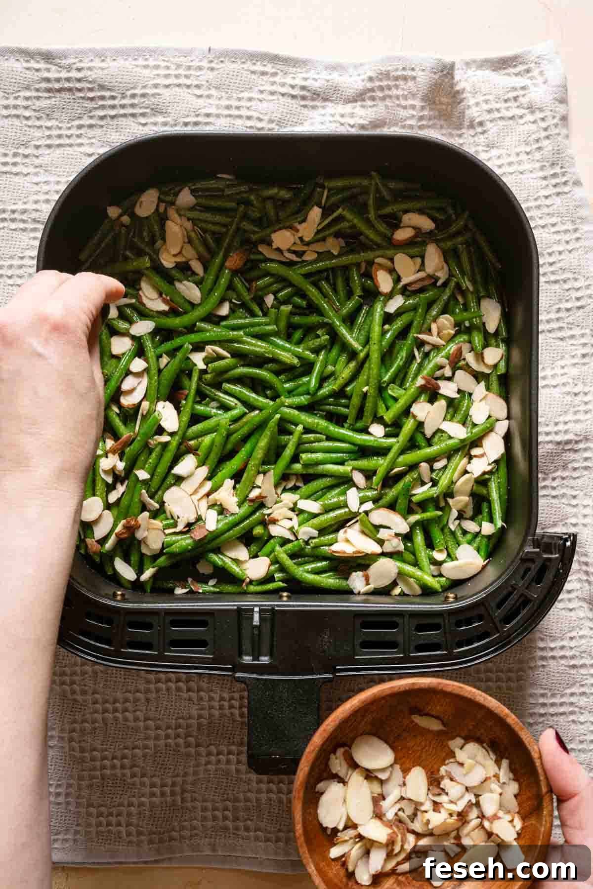 sliced almonds being sprinkled over partially cooked green beans in air fryer basket.
