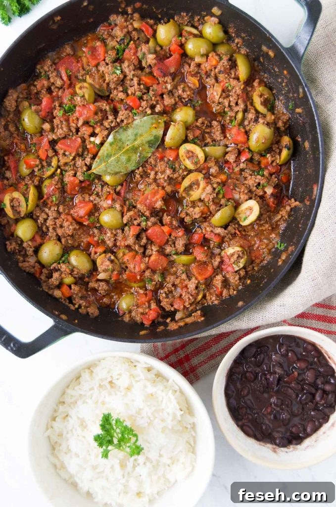 Brazilian picadillo in a black braising pan sits next to a white bowl of rice and a small white bowl holding black beans