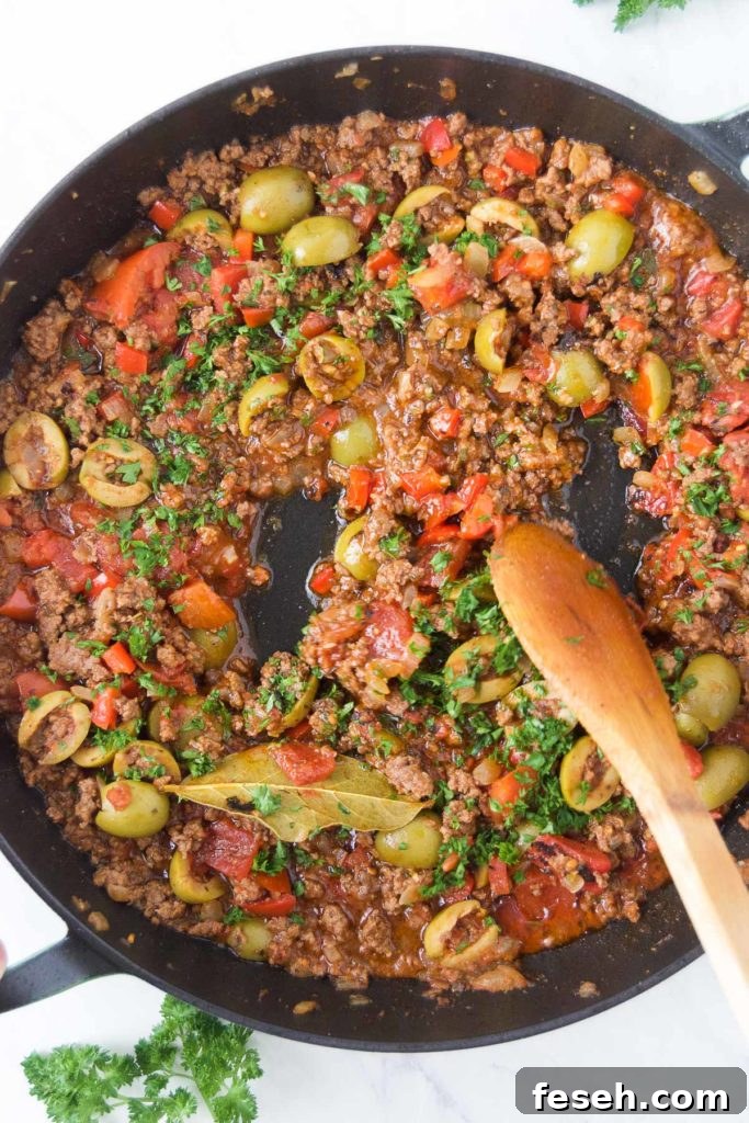 A wooden spoon stirs fresh parsley into a picadillo in a black skillet