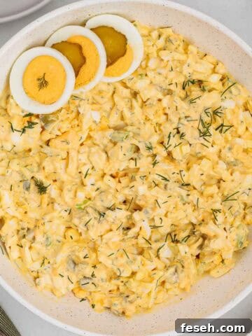 Close up of dill pickle egg salad in a serving bowl topped with sliced hard boiled eggs and sprinkled with fresh dill.