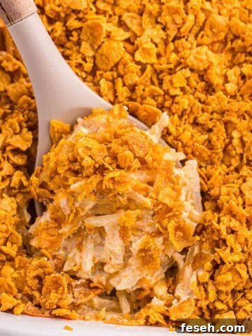 Serving spoon scooping baked hash brown casserole from a baking dish, showing the creamy interior, shredded potatoes, and crisp golden corn flake topping.