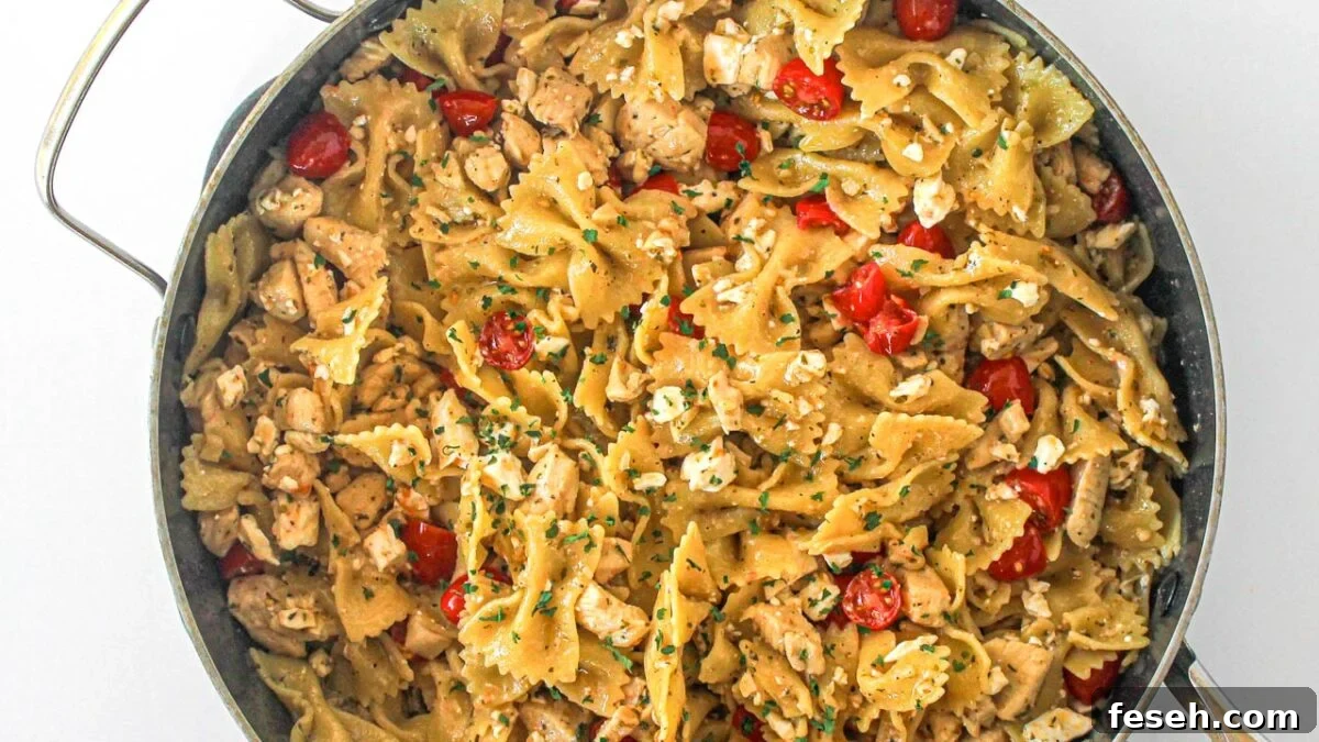 A skillet filled with bowtie pasta, diced chicken, cherry tomatoes, crumbled cheese, and herbs.