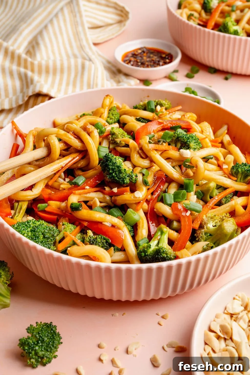 Side-angle image of a bowl of sesame udon noodles with broccoli, carrots, and red bell peppers. Chopsticks lift noodles, with a striped kitchen towel and bowls of chili crisp and chopped peanuts in the background.