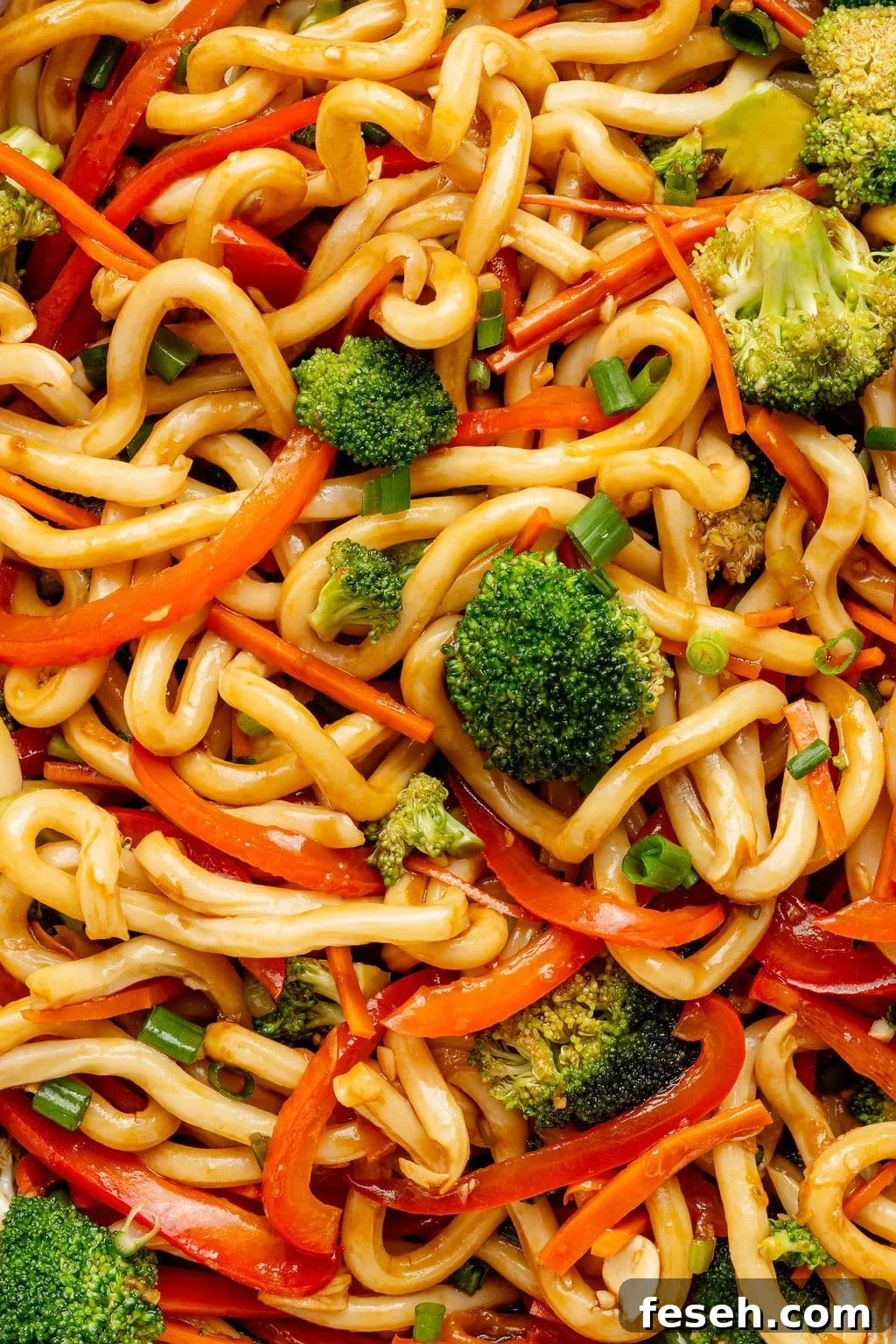Close-up image of glossy udon noodles coated in sesame sauce with broccoli florets, thinly sliced carrots, red bell peppers, and green onions.