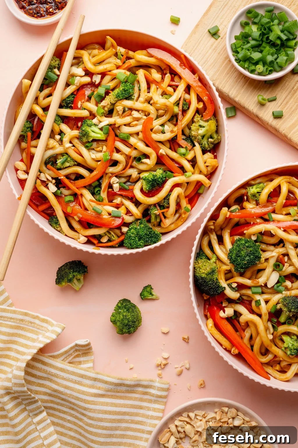 Overhead image of two bowls of sesame udon noodles tossed with broccoli, sliced red bell peppers, and carrots, garnished with green onions and chopped peanuts.