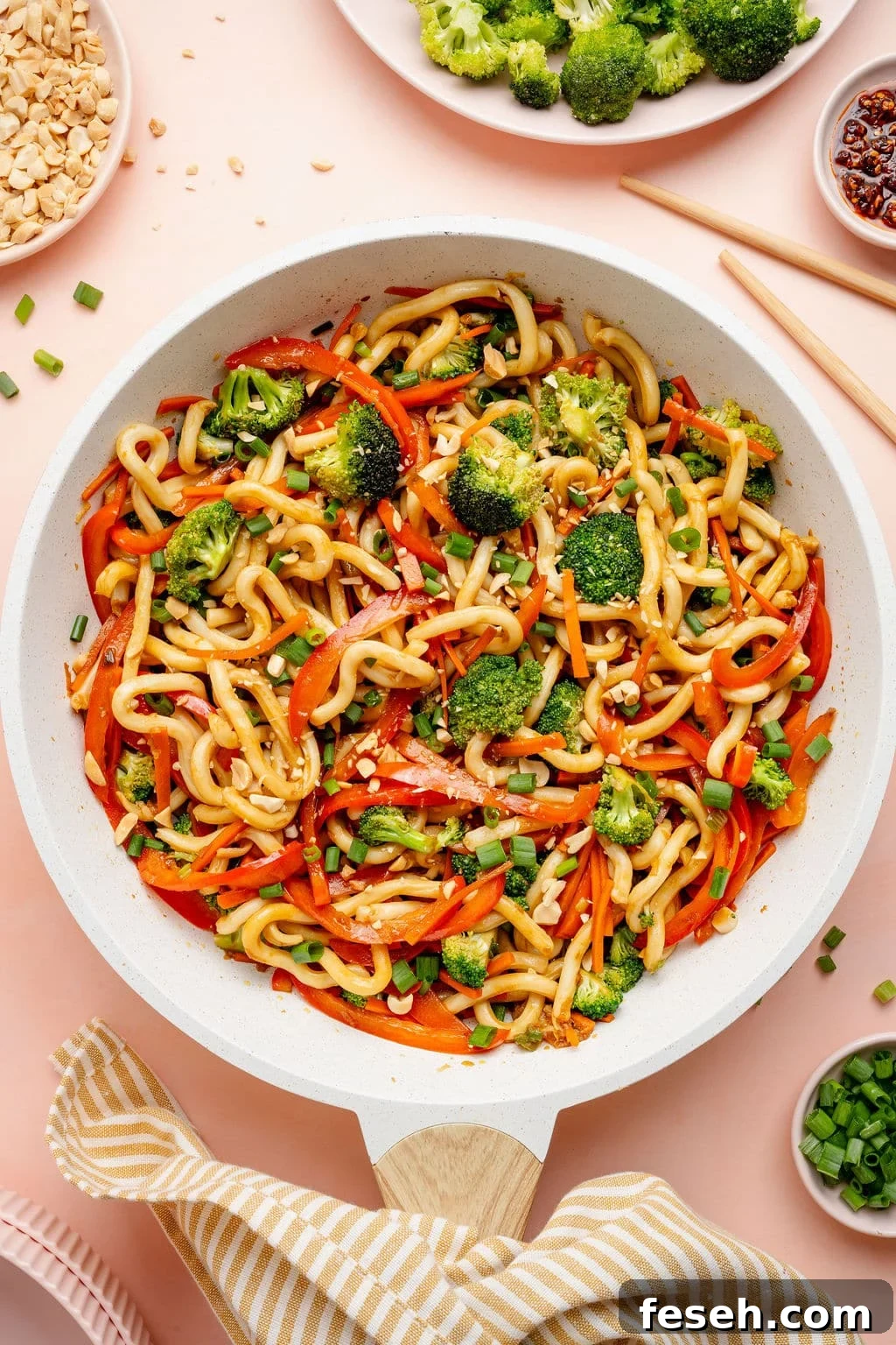 Overhead image of a skillet filled with sesame udon noodles tossed with broccoli, sliced red bell peppers, carrots, and green onions, garnished with chopped peanuts.