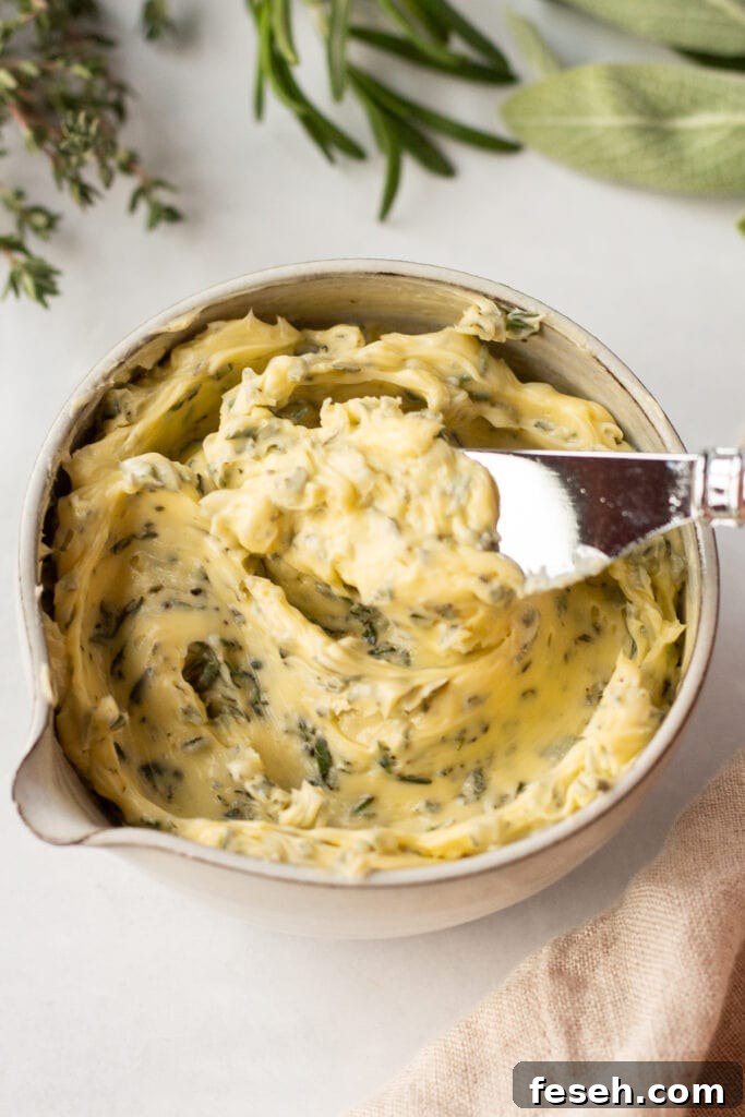 A knife dipping into a small bowl of herb butter that's surrounded by fresh sage, rosemary, and thyme.