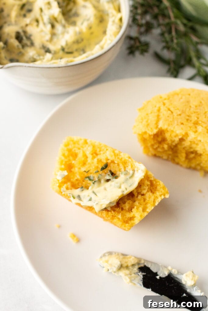 A halved cornbread muffin with a swipe of herb butter on it on a small white plate. A butter knife rests next to it and a small bowl with herb butter, surrounded by fresh herbs, is behind the plate.