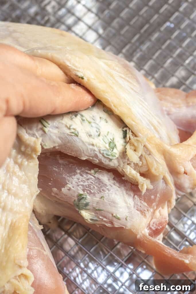 a hand spreading herb butter onto a raw turkey breast.