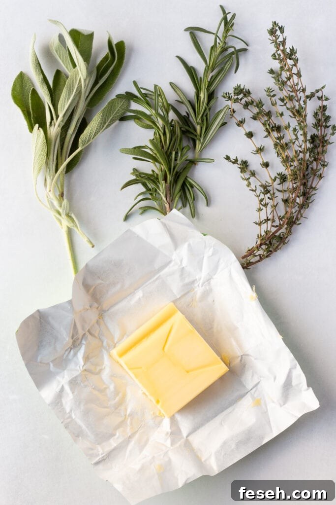 Top down shot of fresh sage, rosemary, and thyme with a slab of butter on a wrapper, all on a white background.