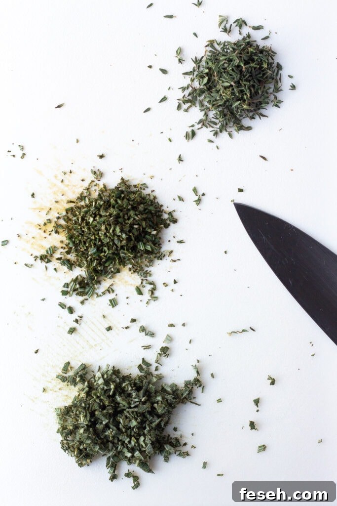 Finely chopped fresh sage, rosemary, and thyme on a white cutting board.