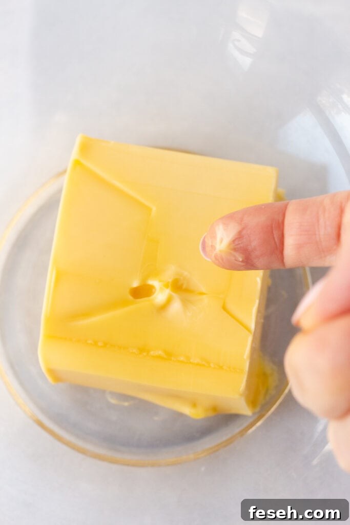 A finger coming up from softened butter in a clear bowl.