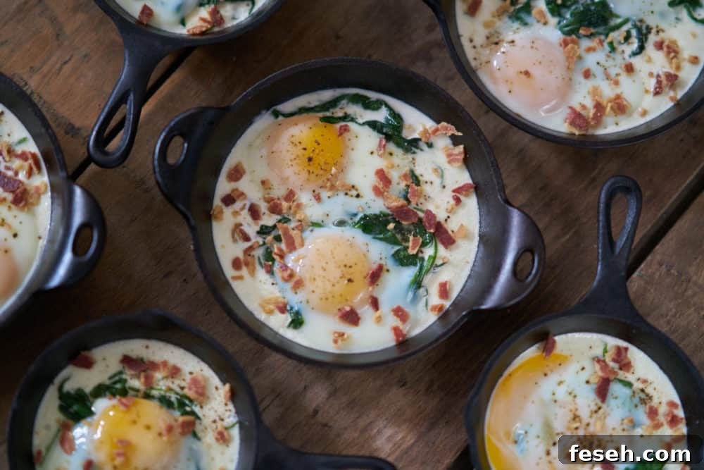Several servings of eggs en cocotte with spinach and bacon in small cast-iron skillets and serving dishes are displayed on a wooden surface. 