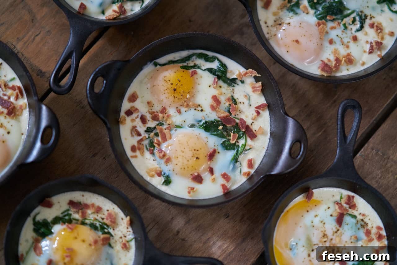 Several servings of eggs en cocotte with spinach and bacon in small cast-iron skillets and serving dishes are displayed on a wooden surface.