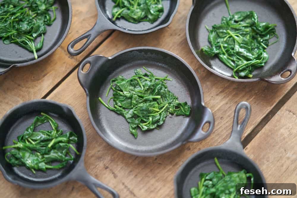 Several small cast-iron skillets and serving dishes containing a small amount of cooked spinach are displayed on a wooden surface.