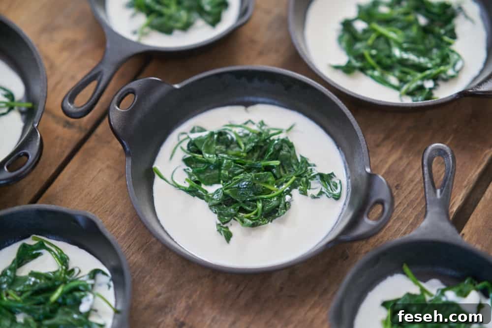 Several small cast-iron skillets and serving dishes containing a small amount of cooked spinach and some heavy cream are displayed on a wooden surface.
