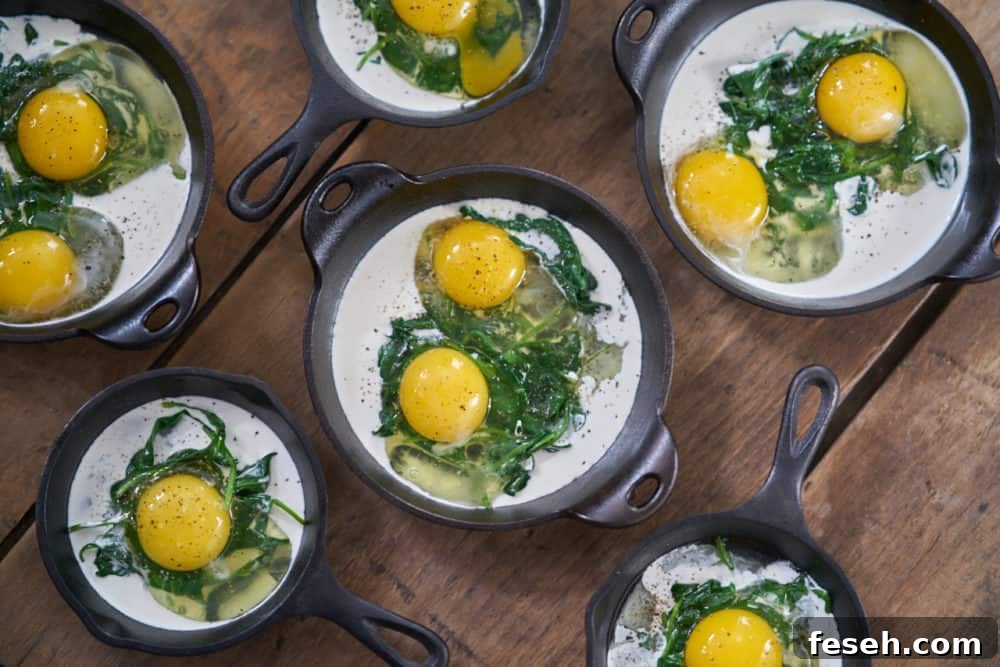 Several small cast-iron skillets and serving dishes containing a small amount of cooked spinach, raw eggs, and some heavy cream are displayed on a wooden surface.