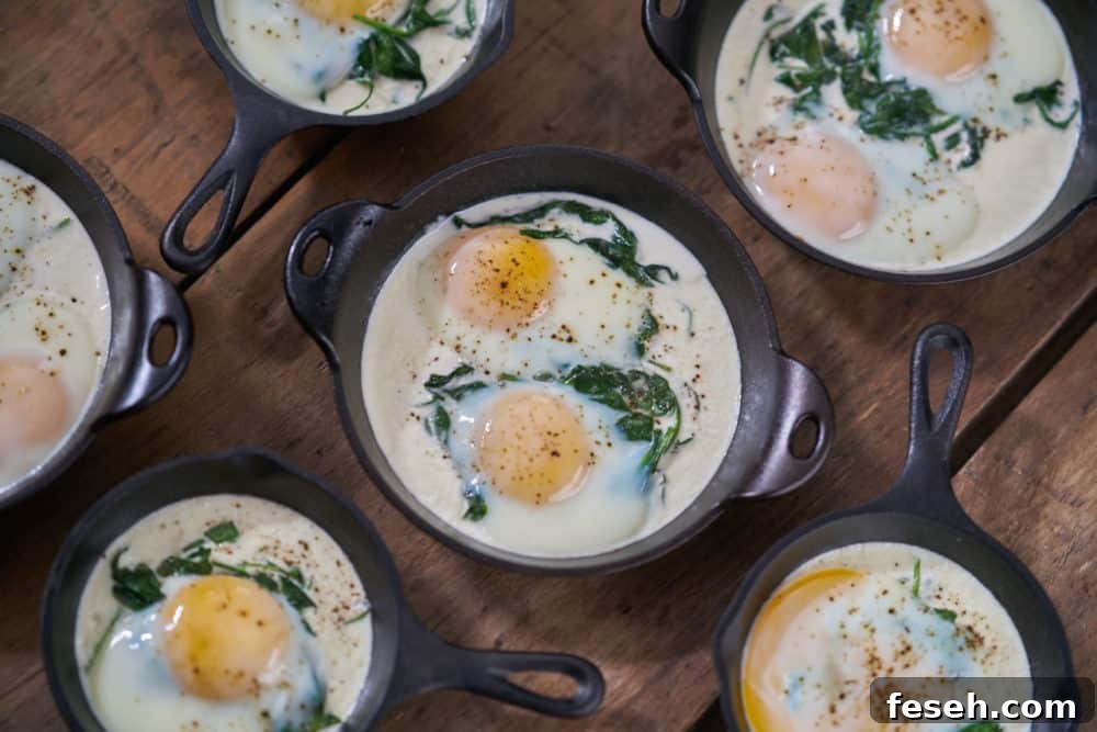 Several servings of eggs en cocotte with spinach in small cast-iron skillets and serving dishes are waiting to be topped with bacon. They are displayed on a wooden surface.
