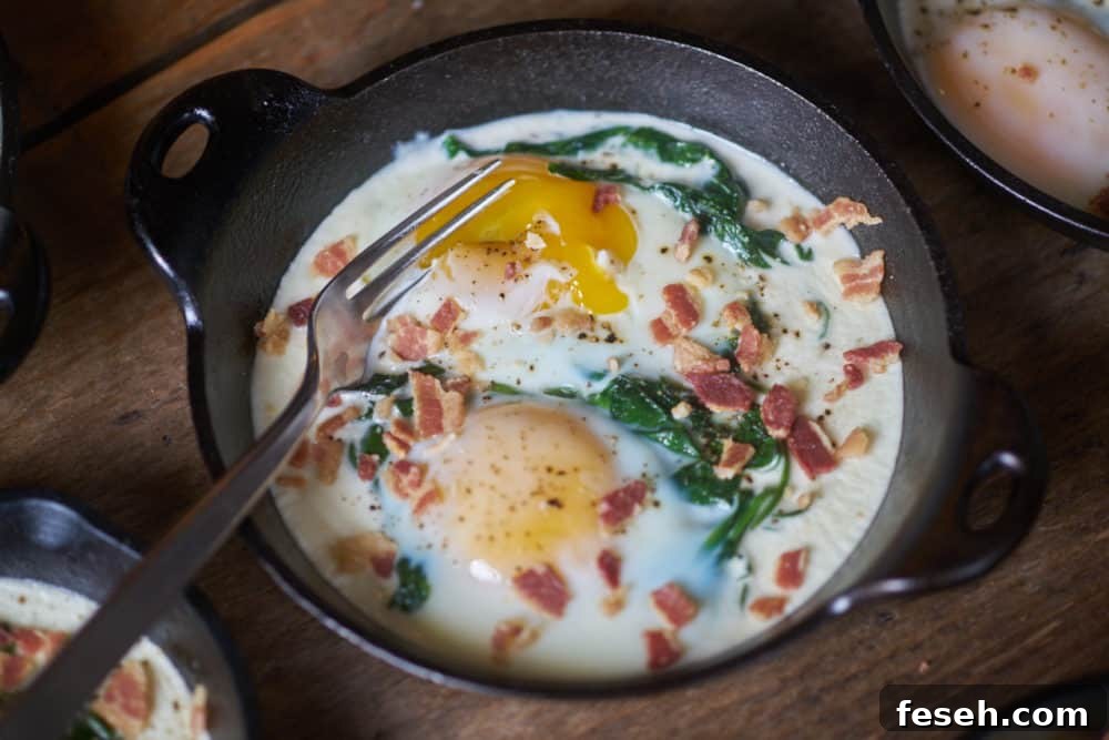Eggs en cocotte with spinach and bacon in a small cast-iron serving dish with a fork that has broken open the egg yolk. The dish is displayed on a wooden surface.