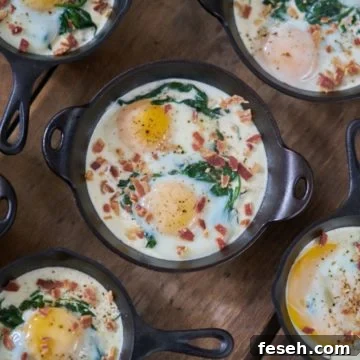 Several servings of eggs en cocotte with spinach and bacon in small cast-iron skillets and serving dishes are displayed on a wooden surface.
