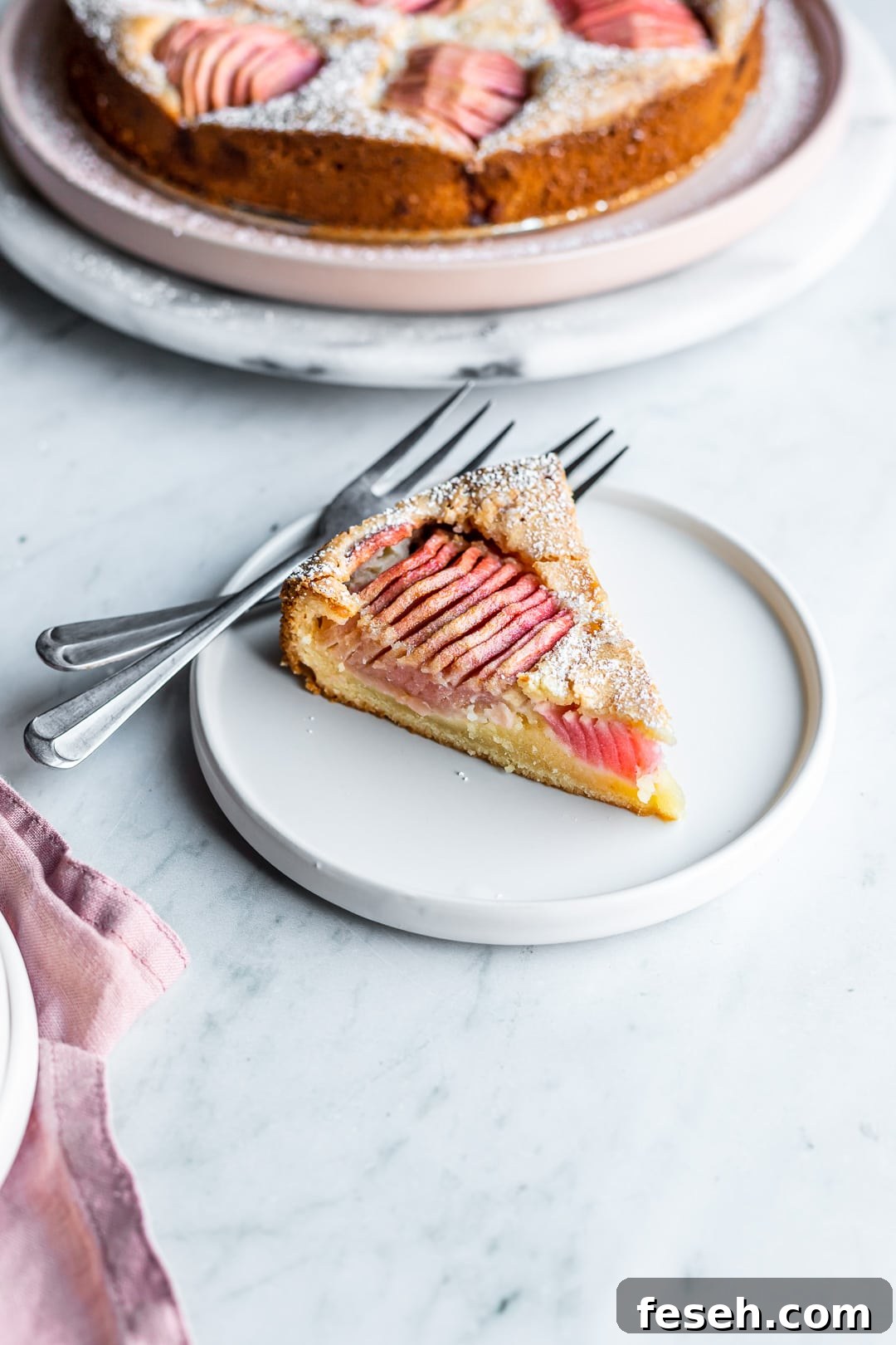 Slice of almond cake with pink apples on a white plate and marble background