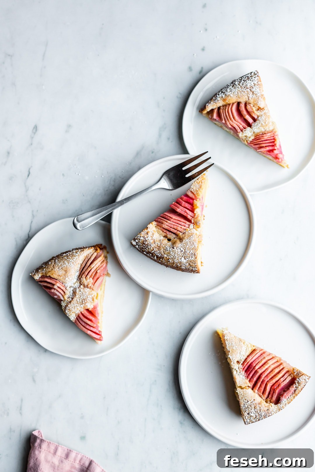 Slices of almond cake with pink apples on four white plates on a marble background
