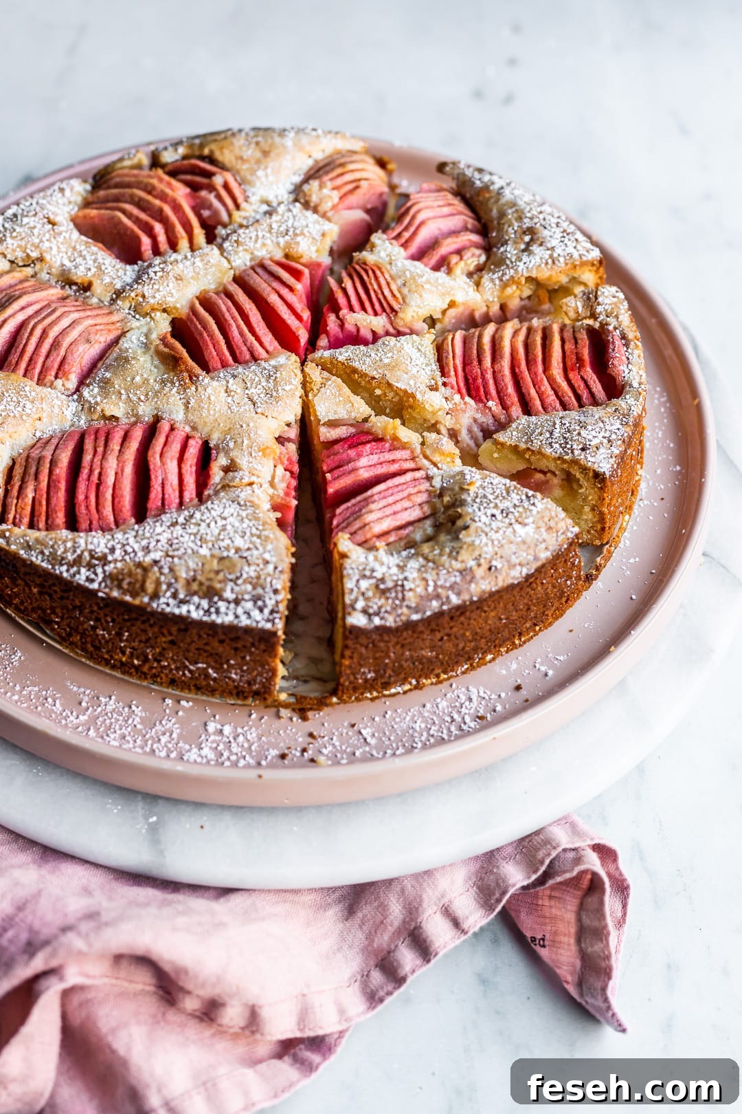 Angled view of sliced almond cake with pink apples on a pink plate and marble background