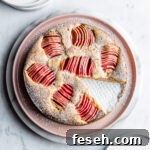 A round cake dusted with powdered sugar has a slice cut out of it. It sits on a pink plate atop a white marble platter, which rests on a white marble surface. There are white plates at the top and bottom of the frame.