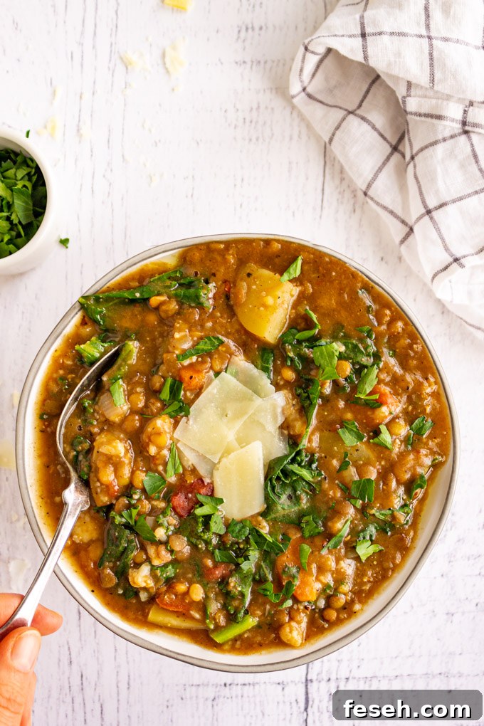 An overhead shot of a bowl of lentil soup with a hand holding a spoon, on a white wood background.