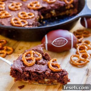 A slice of brownies topped with pretzels next to a cast iron skillet and a little football.