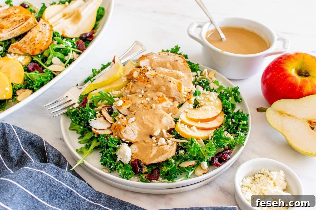 An autumn salad with chicken features sliced chicken, kale, and apple slices, topped with a creamy dressing. A bowl of dressing, pear, apple, and blue cheese crumbles are placed near the salad.