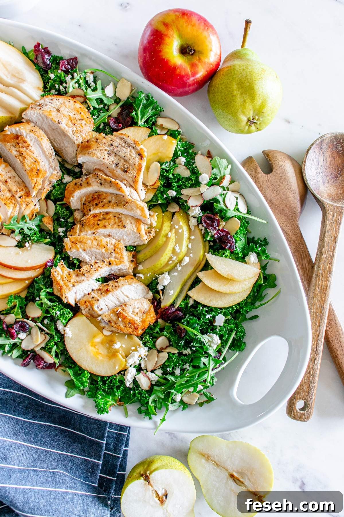 An autumn salad with chicken featuring sliced grilled chicken, kale, apple and pear slices, almonds, cranberries, and crumbled cheese is displayed on a white platter with wooden serving utensils and whole fruits nearby.