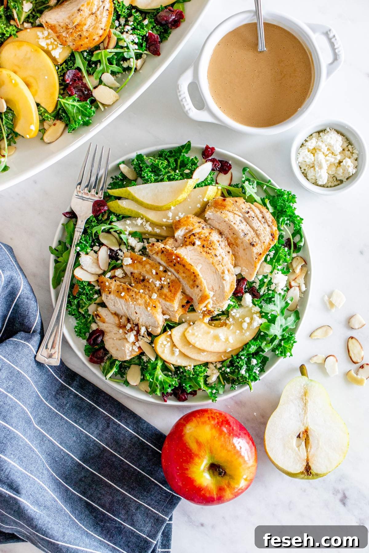 A plate with grilled chicken salad, containing kale, pear slices, apple slices, cranberries, almonds, and cheese crumbles. A small bowl of dressing and a fork are next to the plate. An apple, pear, and almonds beside the plate complete this autumn salad with chicken.